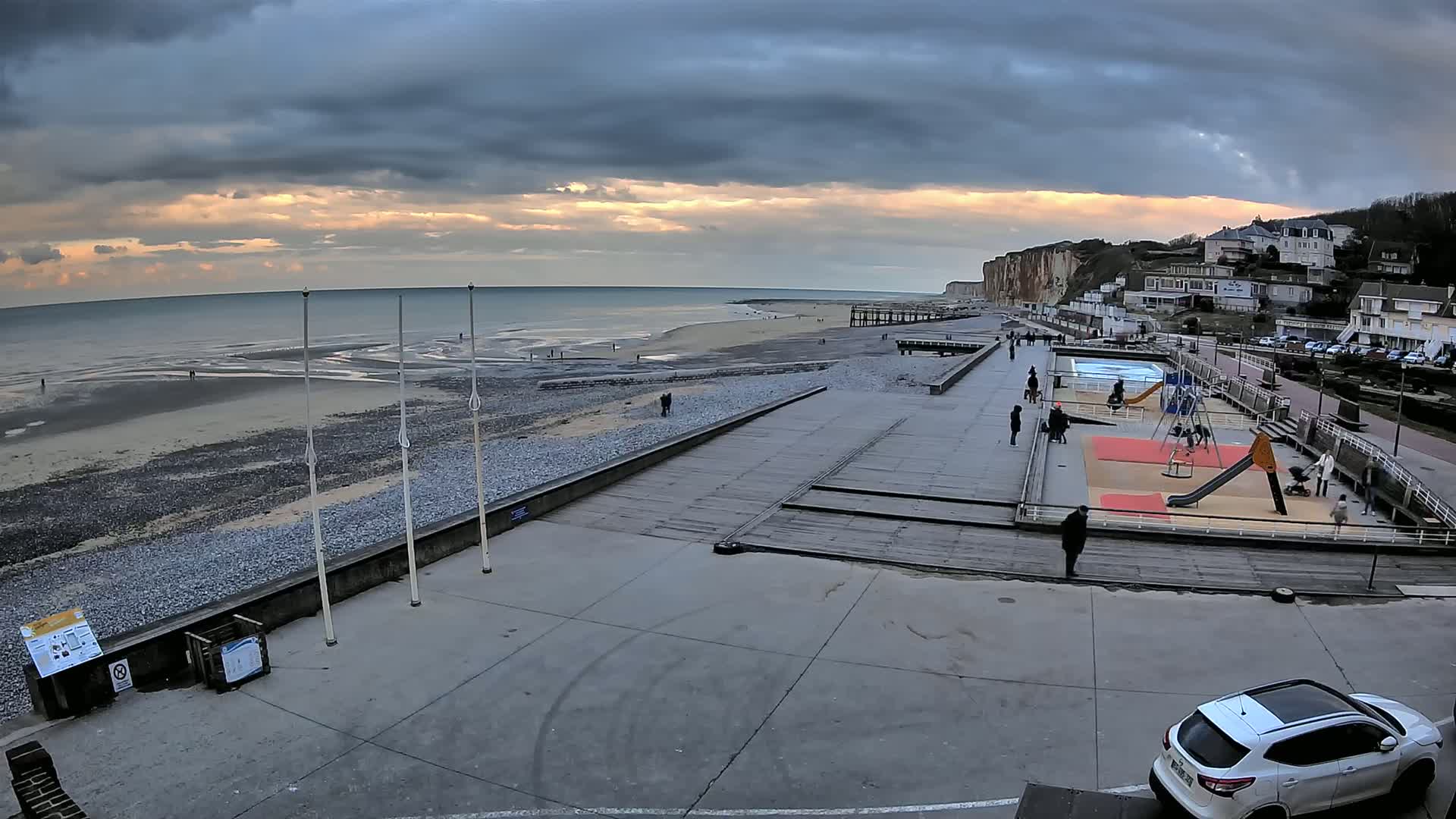 A mostly cloudy sunset illuminates a beach with a boardwalk, playground, and residential buildings overlooking the ocean.