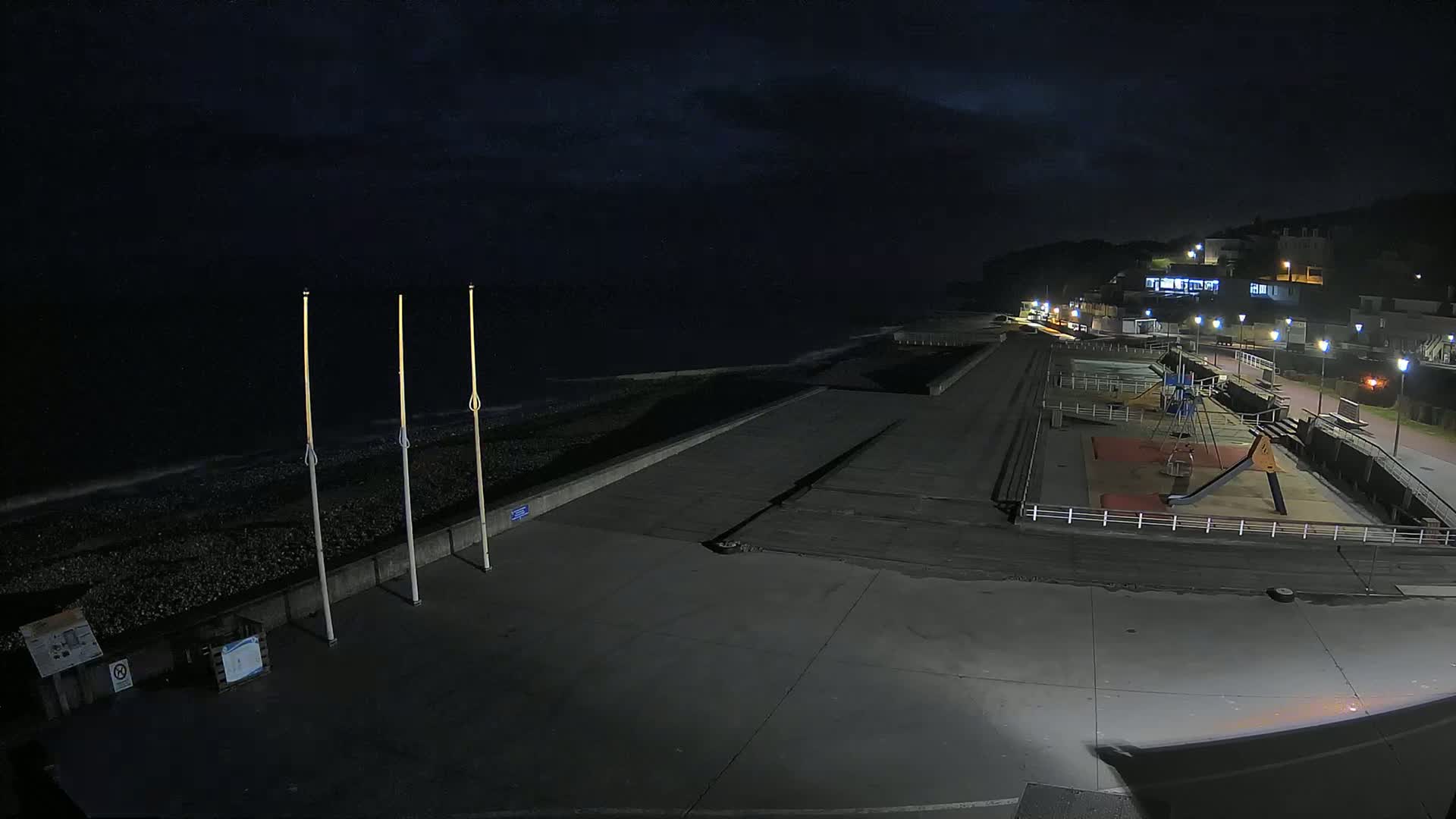 A nighttime view of a mostly empty beach promenade with a playground, a pebble beach, and buildings on a hill in the background under a dark, cloudy sky.