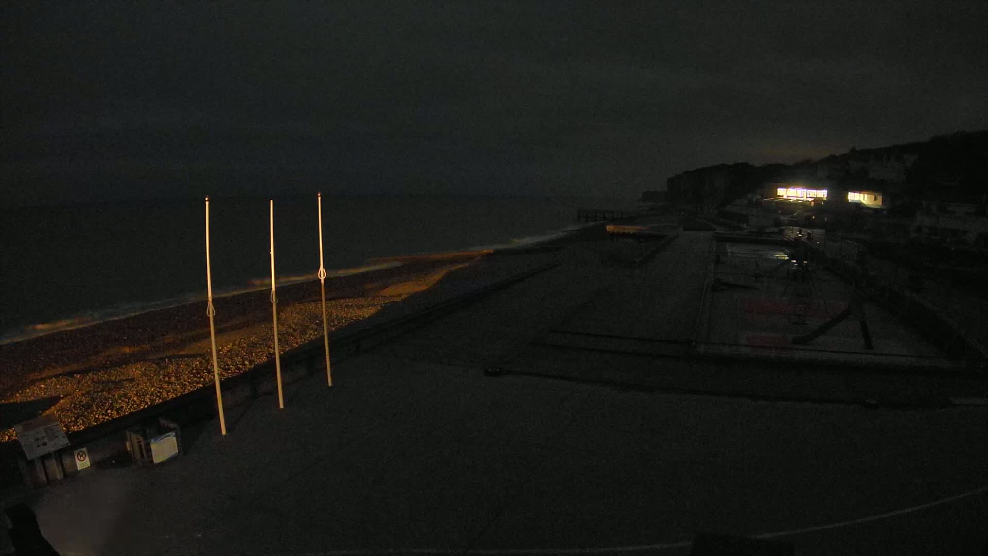A dark, nighttime beach scene shows a pebble shore with calm waves, three tall poles, and a building with illuminated windows in the distance under a mostly cloudy sky.