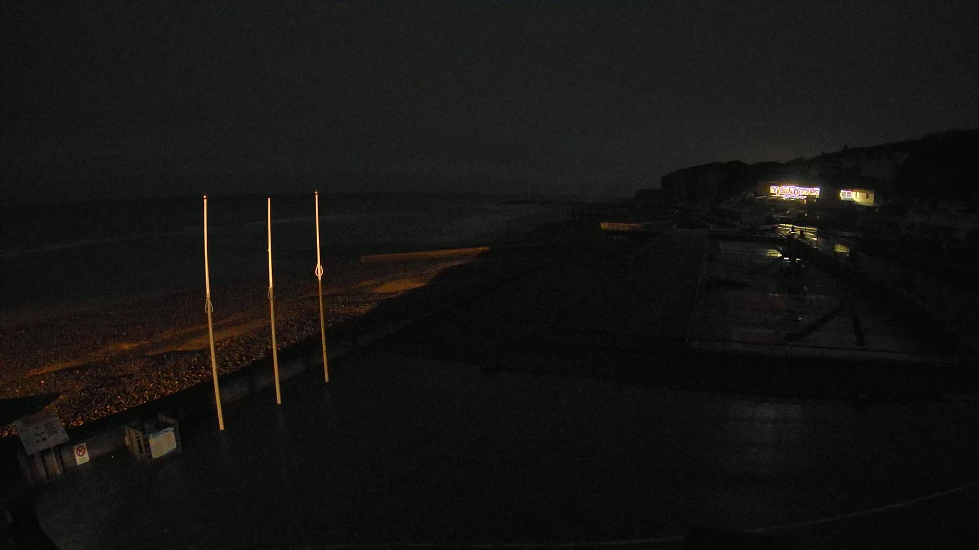 Three tall, thin poles stand on a dark beach at night near a dimly lit building under a cloudy sky.
