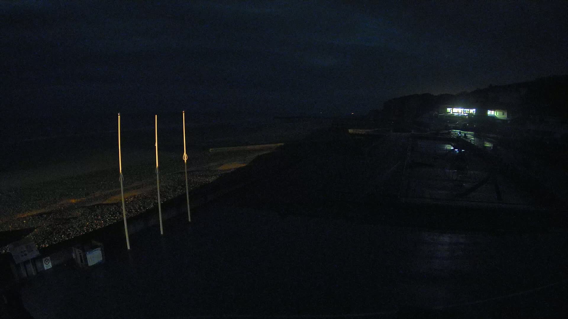 Three illuminated poles stand on a dark beach at night near a dimly lit building.