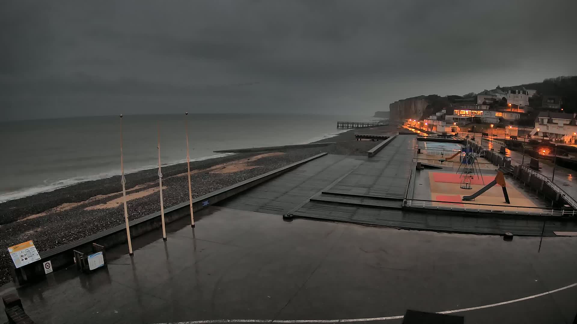 A wet, dark gray coastal scene shows a beach with a playground, a pier, and buildings under an overcast sky.