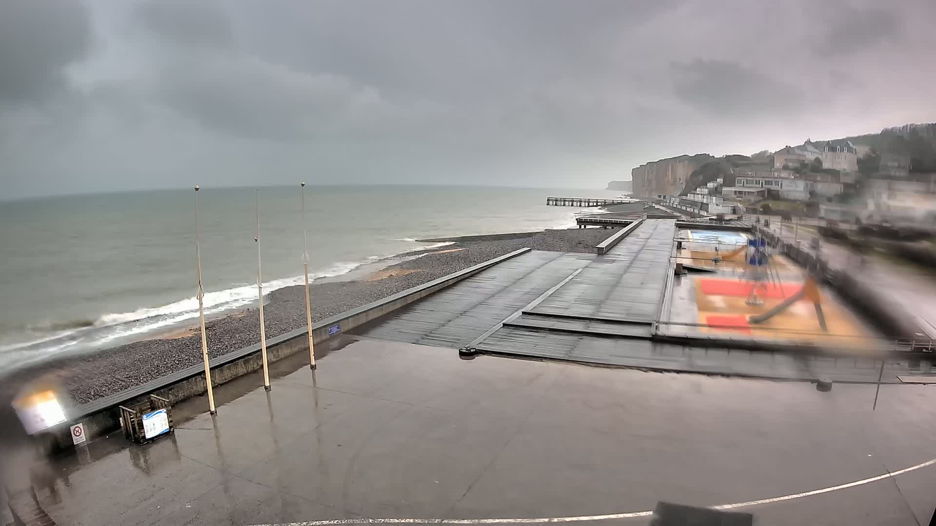 A rain-soaked coastal promenade features a playground and a pier extending towards a grey, overcast sea under a stormy sky.