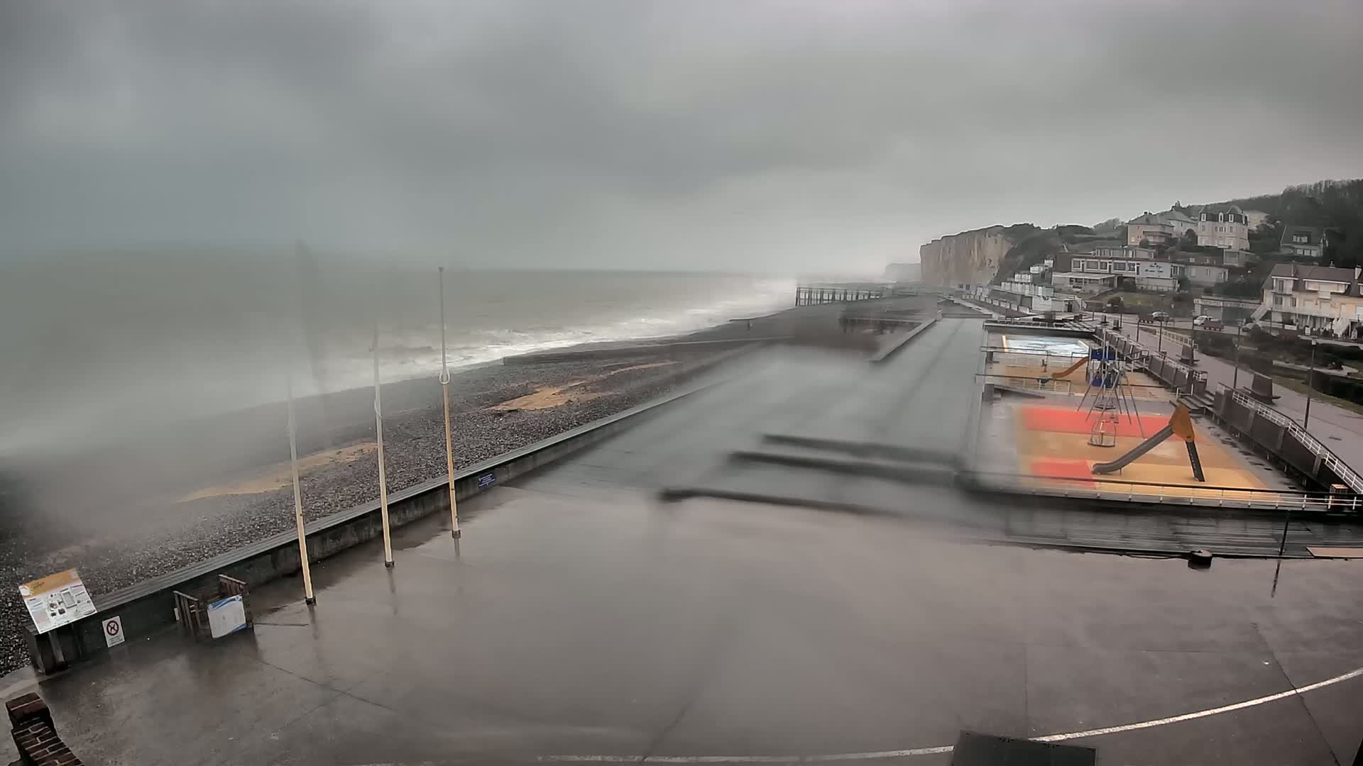 A rain-swept seaside promenade shows stormy waves crashing against a pebble beach, with a children's playground and buildings visible in the background.