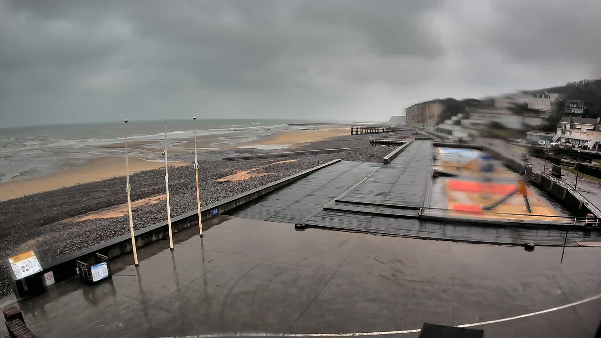 A rain-soaked beach and promenade scene shows a grey, overcast sky, a dark, choppy ocean, and a rocky shoreline with a boardwalk and steps leading down to the water.