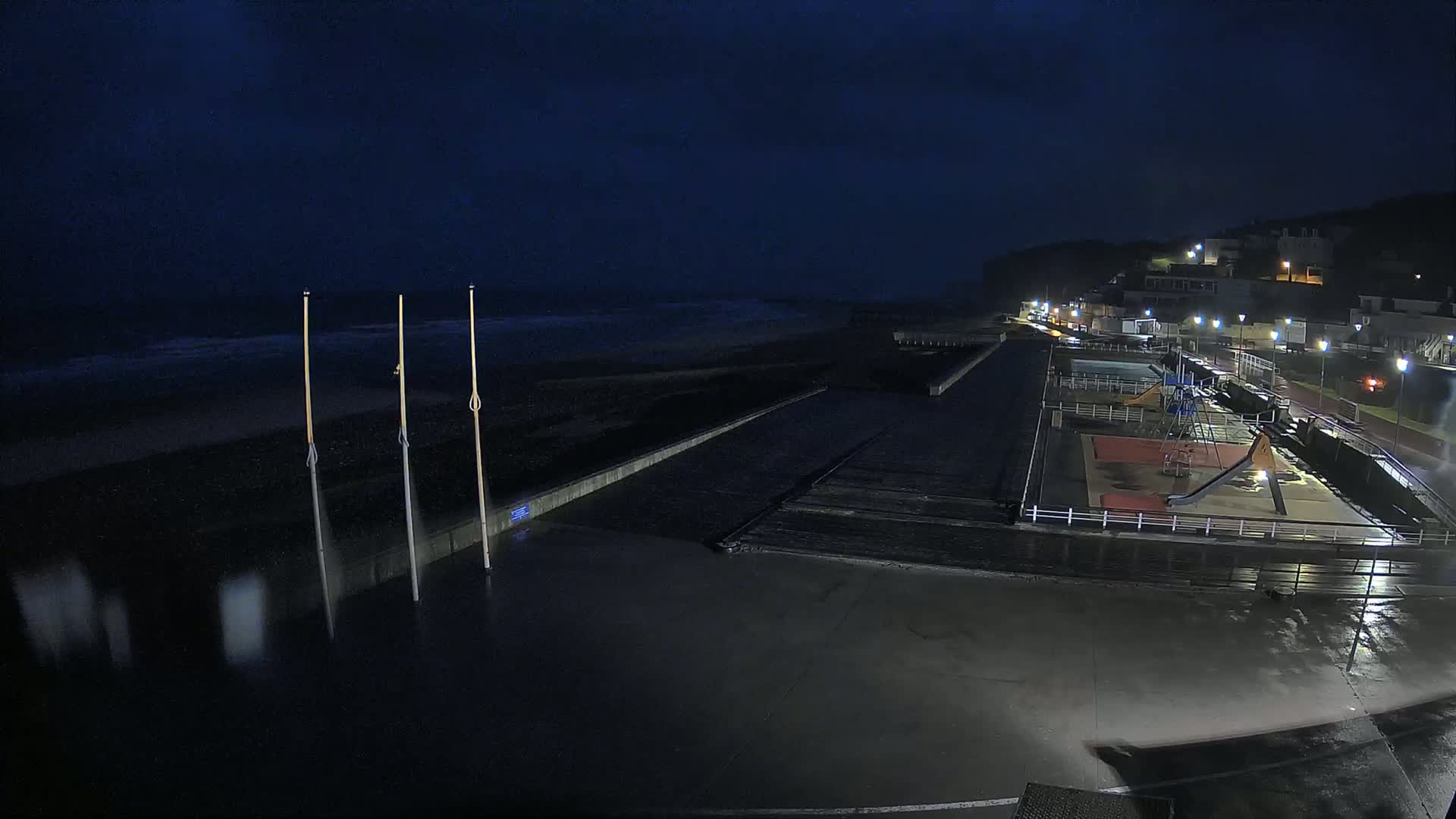 A dark, nighttime seaside scene shows a mostly empty playground next to a wet, dark beach under a cloudy sky.