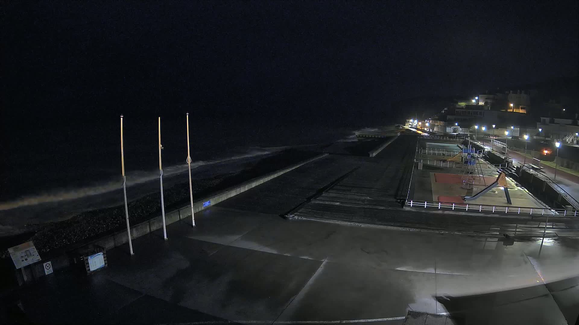 At night, under dark skies, a mostly empty seaside promenade is lit by streetlights, with small waves crashing against a seawall near a playground.