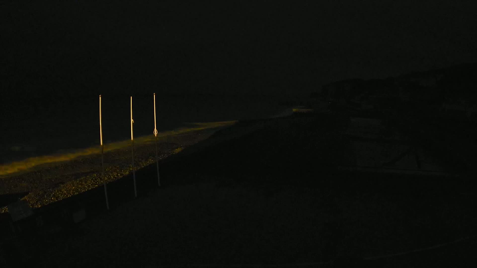 Three tall, thin, illuminated poles stand on a dark beach at night, with faint light reflecting off the water.