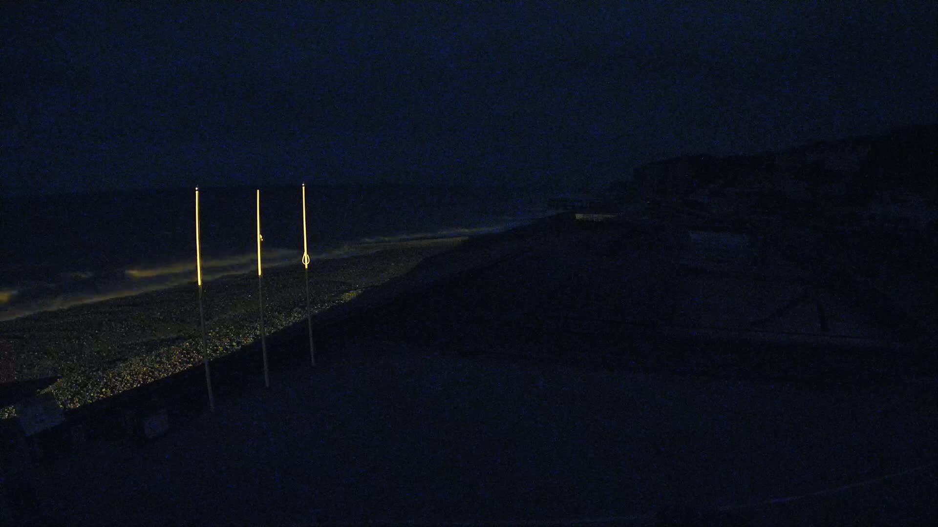 Three tall, slender, illuminated posts stand on a dark, pebble beach at night next to a dark, shadowed cliff under a nearly cloudless sky.