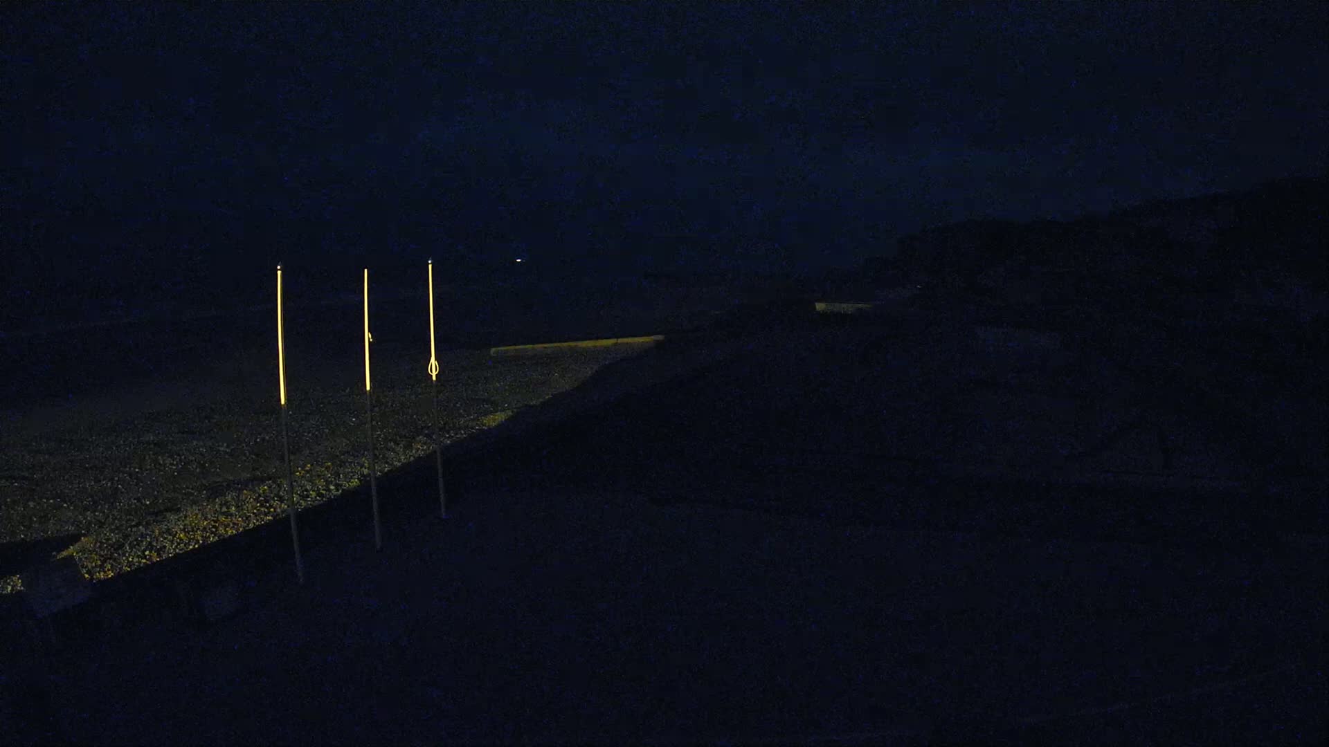 Three tall, thin lights illuminate a dark, pebble-strewn beach at night.