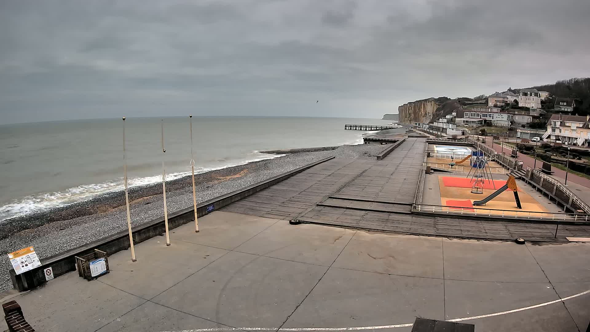 A pebble beach with a wooden boardwalk, playground, and a pier, backed by white buildings and tall cliffs under an overcast sky.