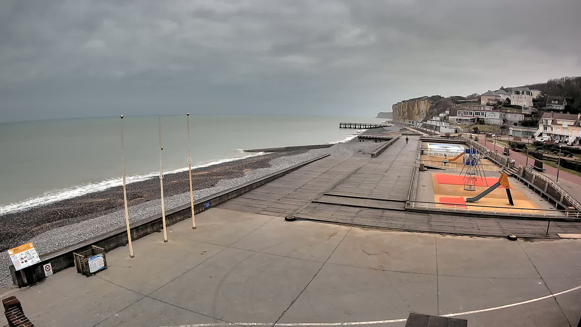 A gray, overcast day shows a pebble beach with a wooden boardwalk, a playground, a pier, and white cliffs in the background.