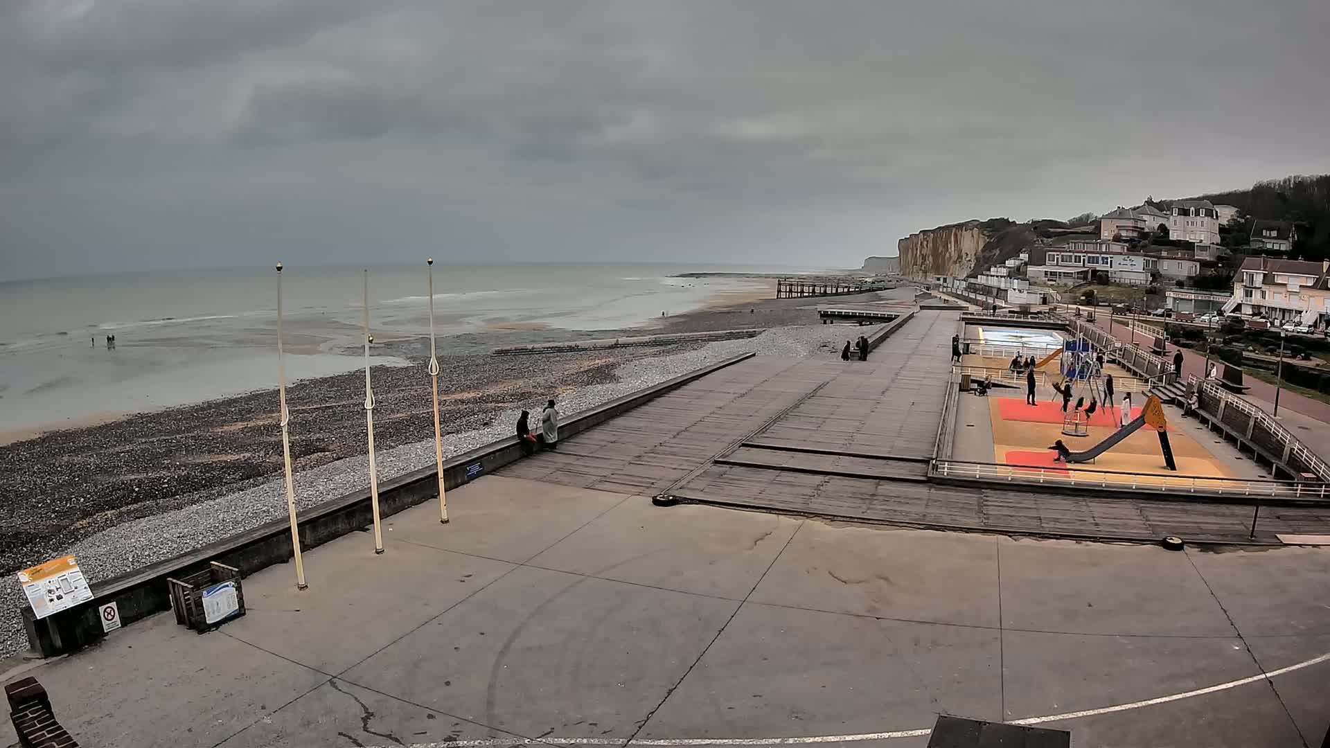 A mostly empty beach and boardwalk overlook a calm sea under an overcast sky, with a playground and a few people visible nearby, and white cliffs in the distance.