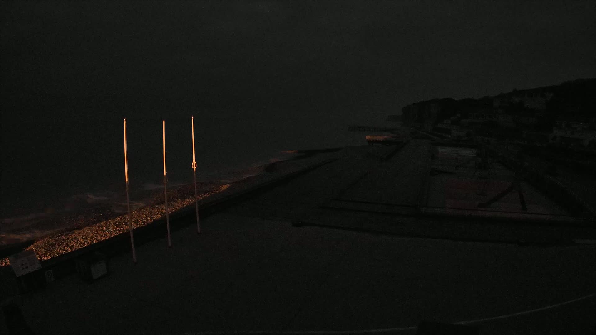 Three tall, slender lights illuminate a dark, pebble-strewn beach under a stormy, overcast sky near a dimly lit coastline.