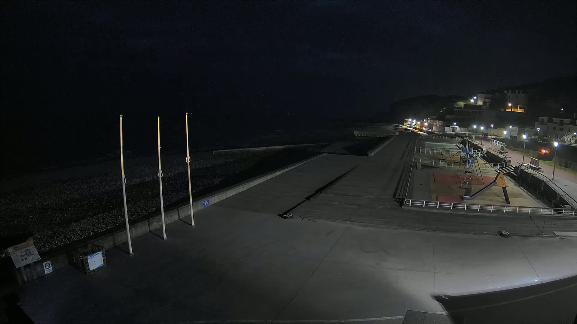 A nighttime view of a mostly empty seaside promenade with a playground and a pebble beach under a dark sky.