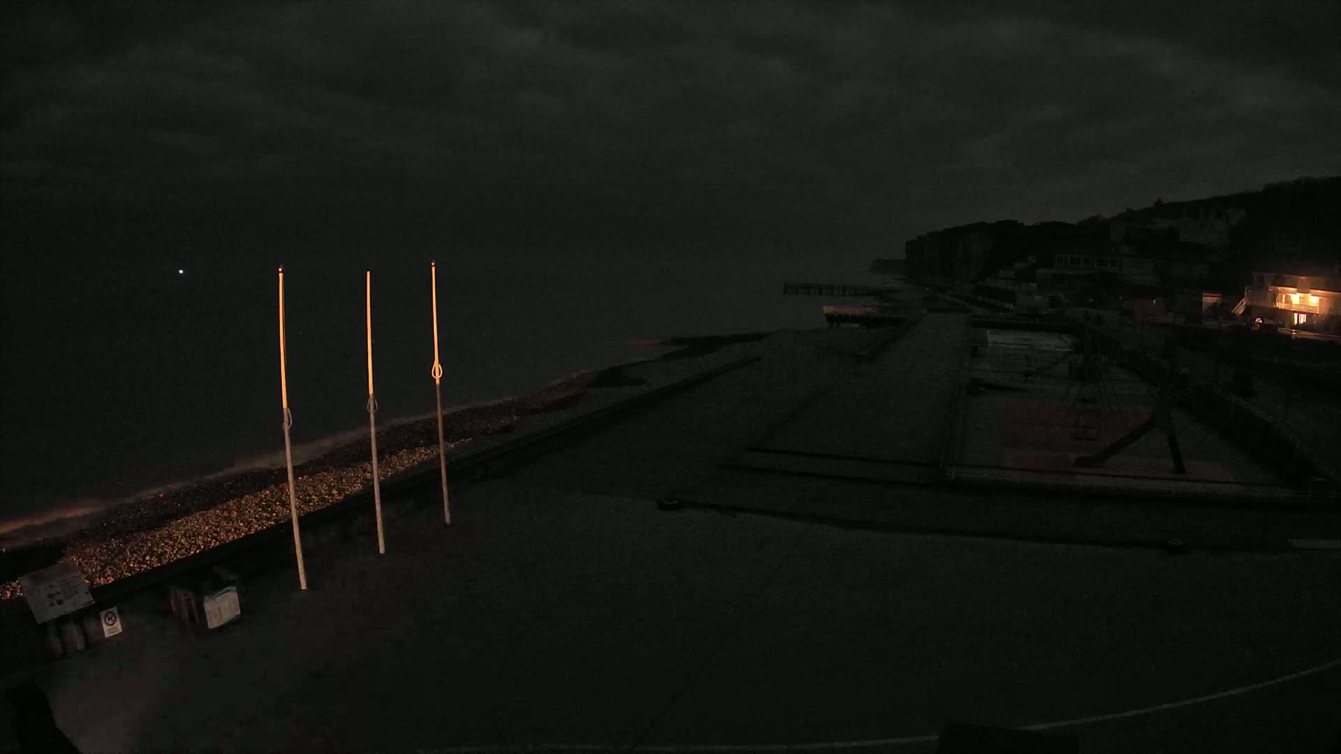 Under a dark, cloudy night sky, a dimly lit seaside area shows a pebble beach, a promenade, and some buildings in the distance.