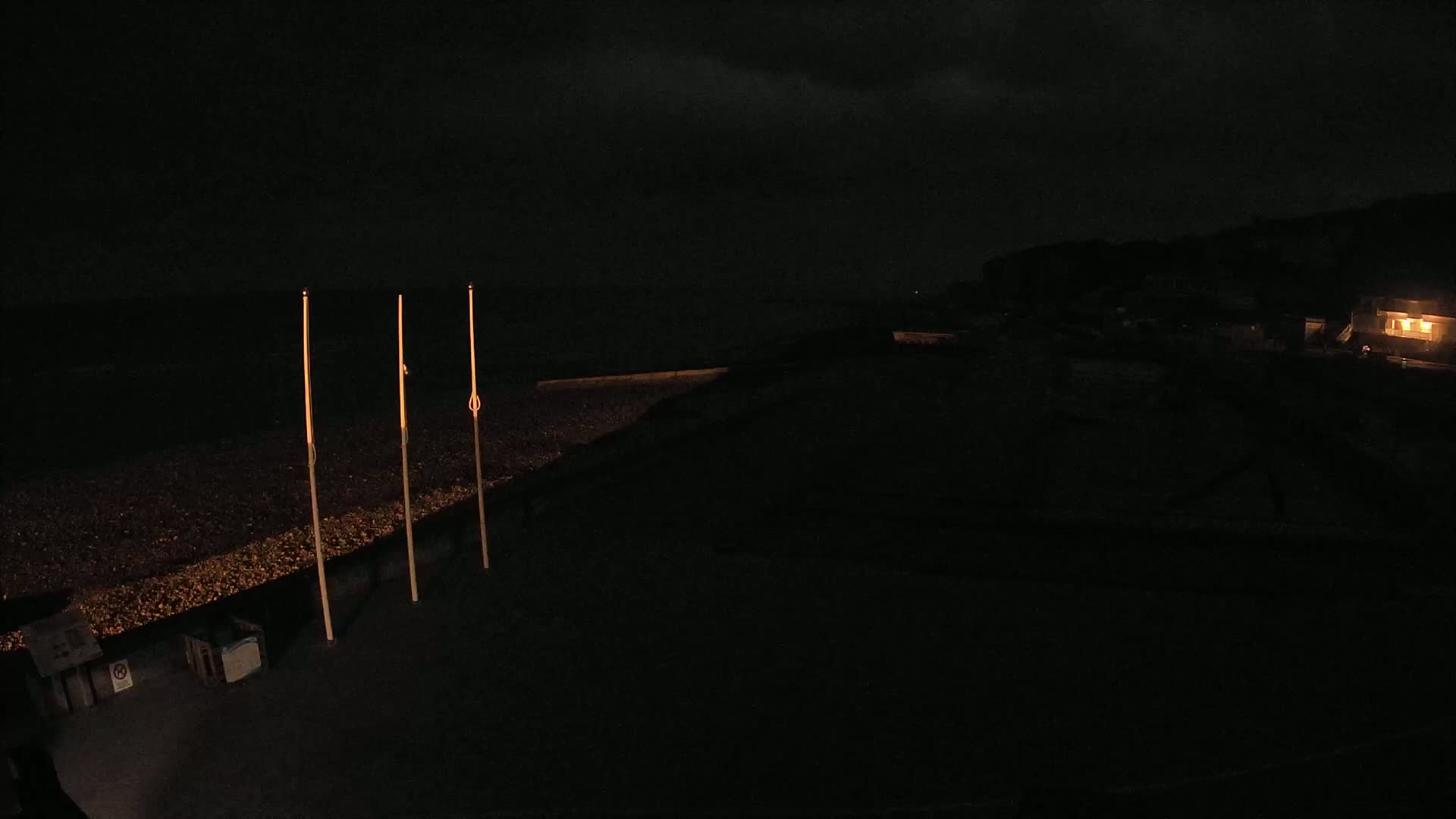 Three tall, slender poles stand on a dark, pebbled beach at night, with a dimly lit building visible in the distance under a cloudy sky.