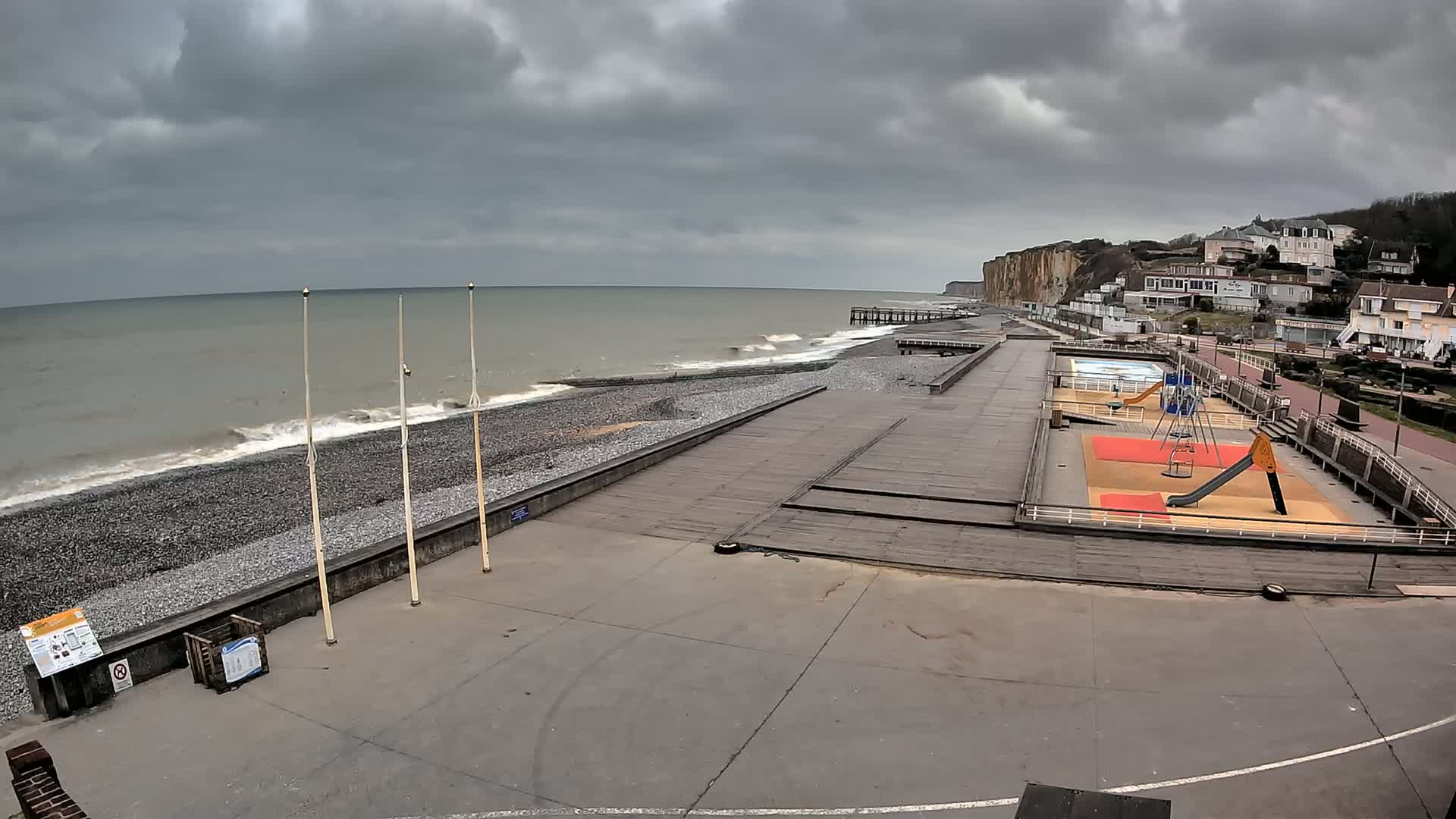 A pebble beach with a playground and wooden boardwalk stretches along a grey, overcast ocean beneath a cloudy sky, backed by white buildings and high cliffs.