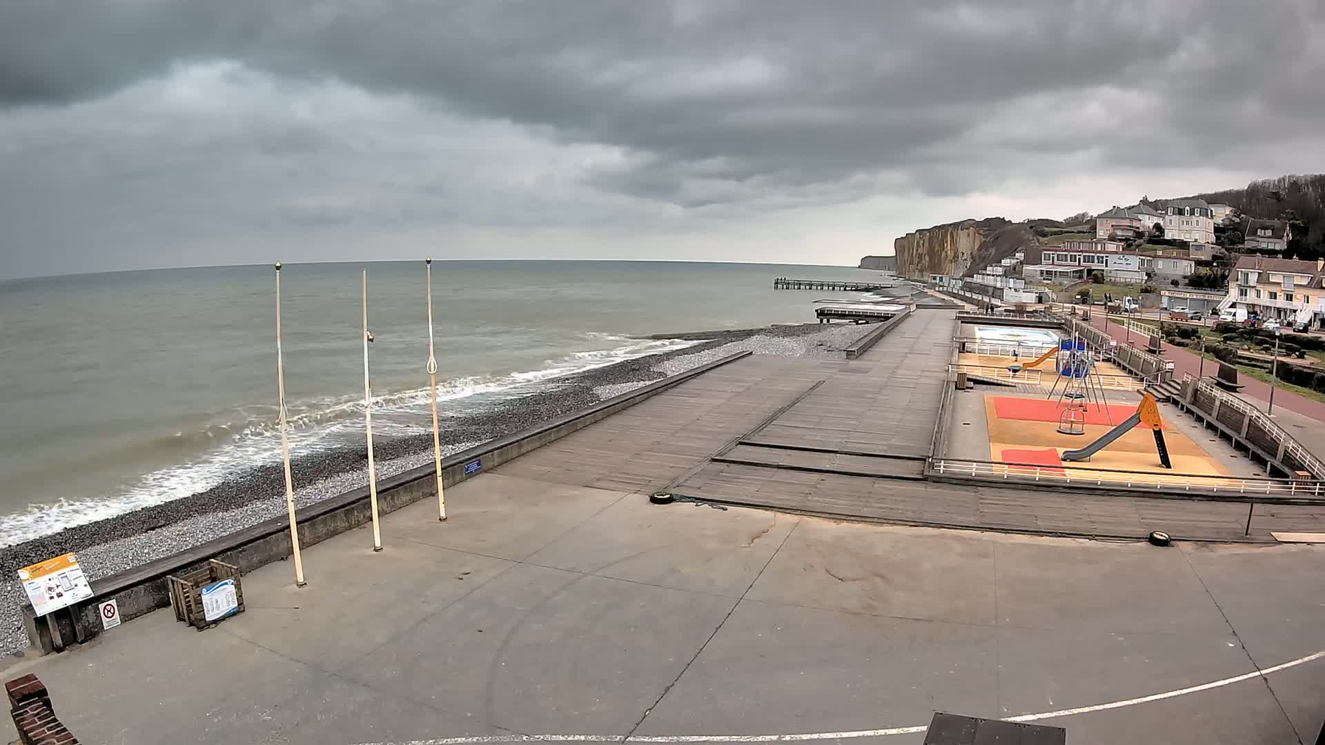 A seaside playground and boardwalk are visible under an overcast sky, with small waves breaking on a pebbled beach near a cliff.