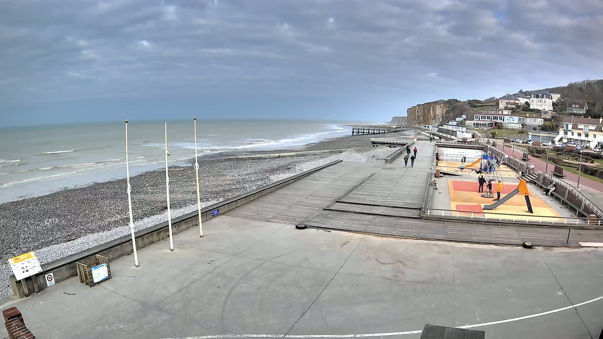 A pebble beach with a wooden boardwalk, playground, and a few people, overlooks a calm sea under a cloudy sky.