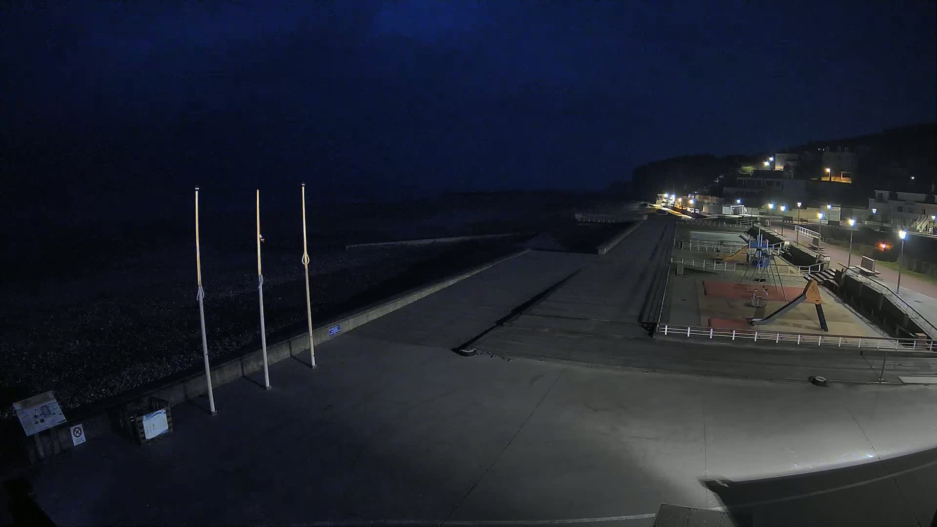 An empty, dark seaside promenade at night features a small playground and a pebble beach under a mostly clear, dark sky.