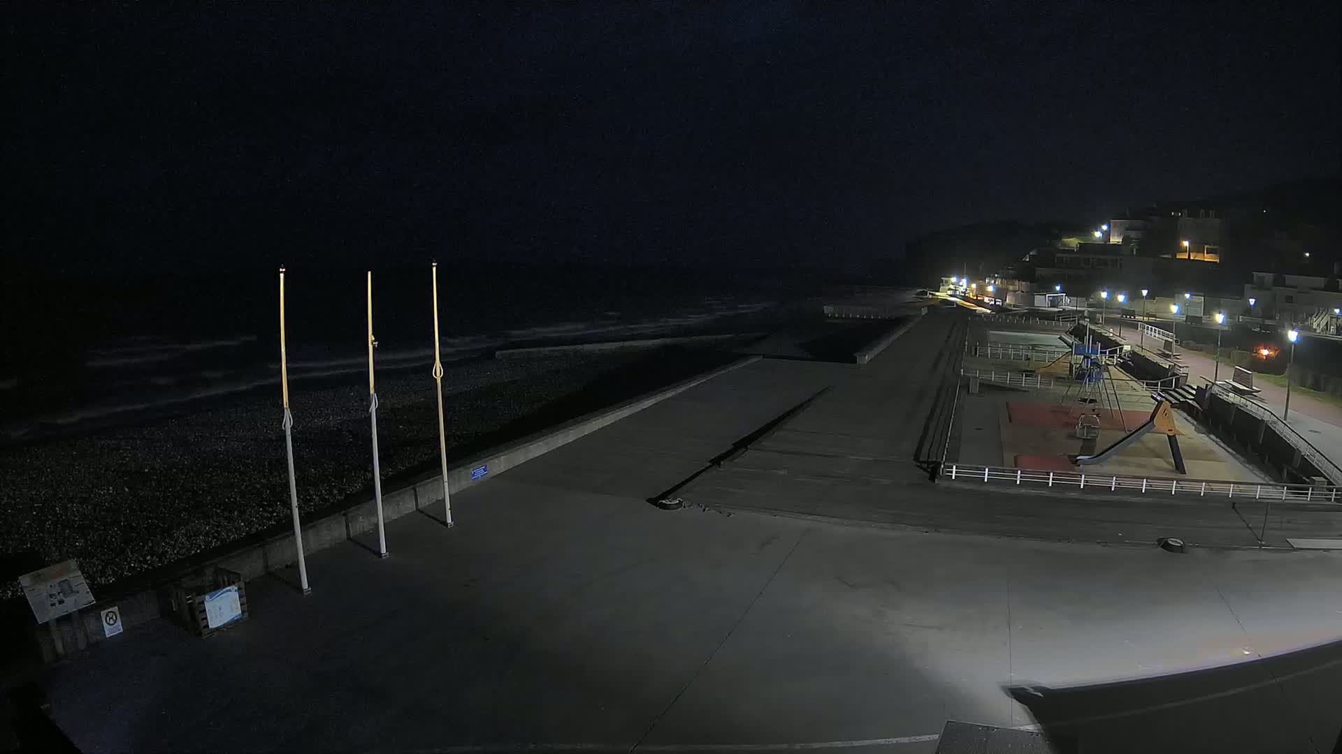 A nighttime view of a mostly empty seaside promenade with a playground, featuring dark, wavy water under a dark sky.