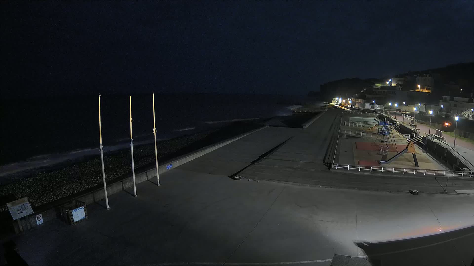 A nighttime coastal scene shows a dark ocean with small waves, a paved walkway next to a pebble beach, a playground with a slide and swing set, and lit buildings on a hillside in the background.