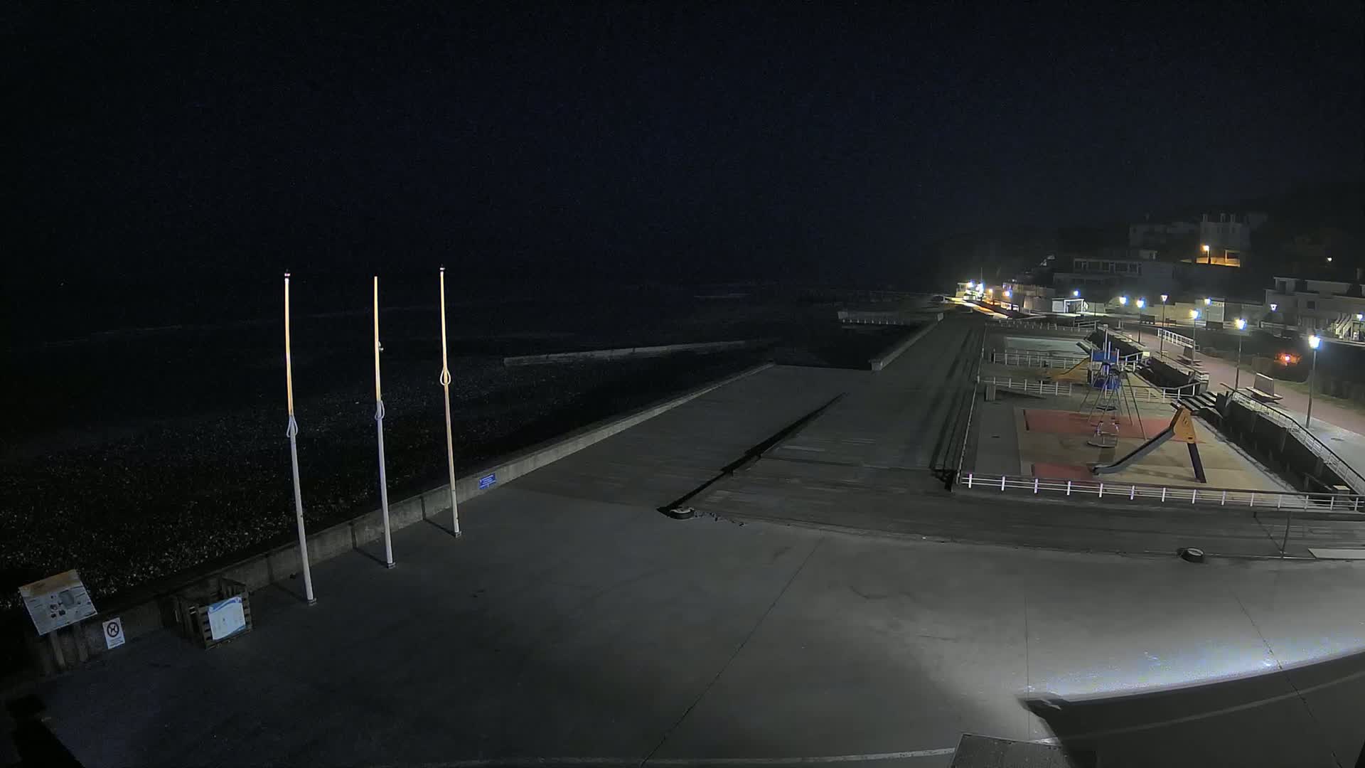 An empty, dark seaside promenade at night, featuring a playground, a sea wall, and some tall poles, with buildings visible in the distance.