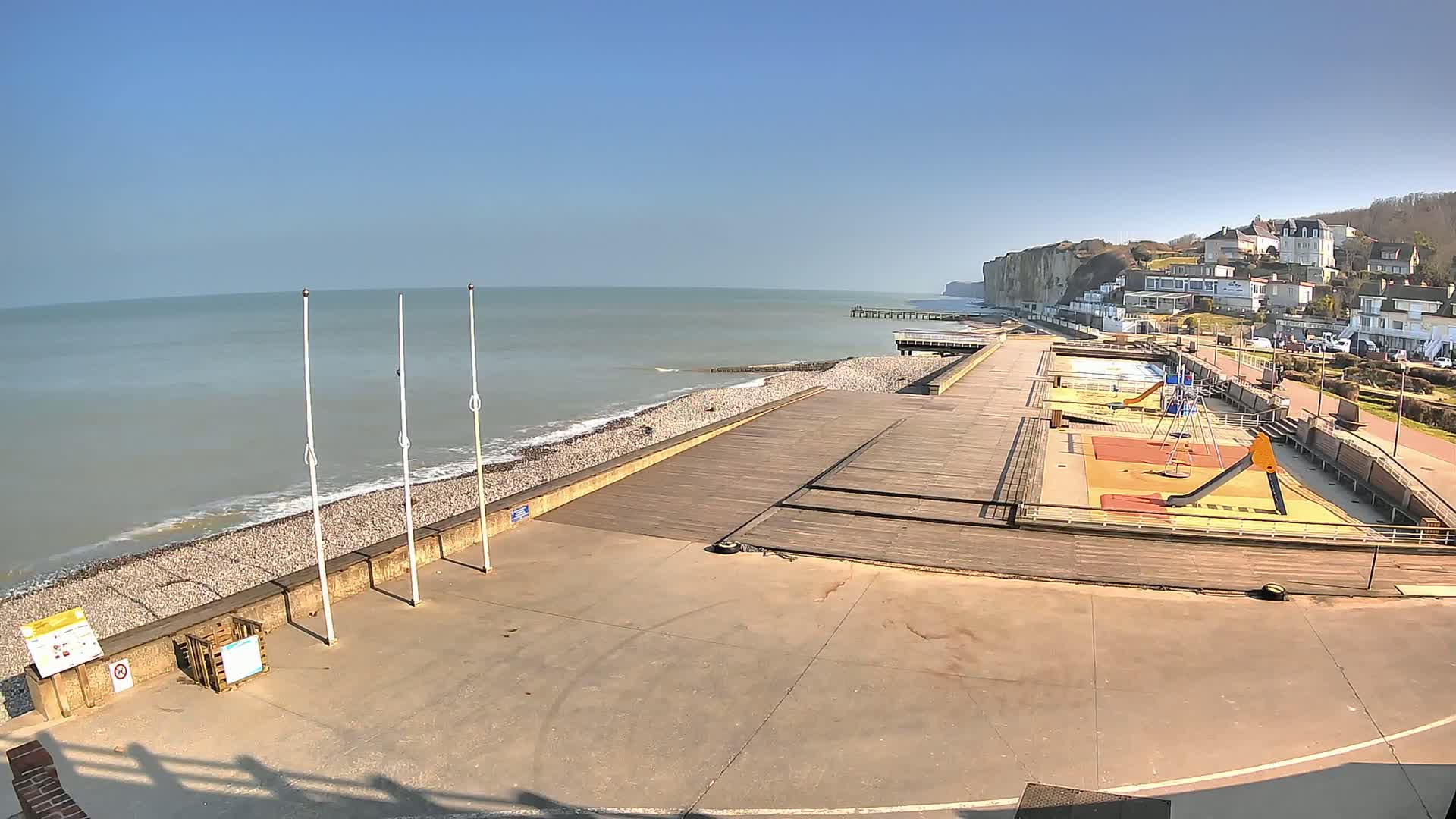 A seaside promenade with a playground and a pier extends along a pebbled beach under a clear blue sky.