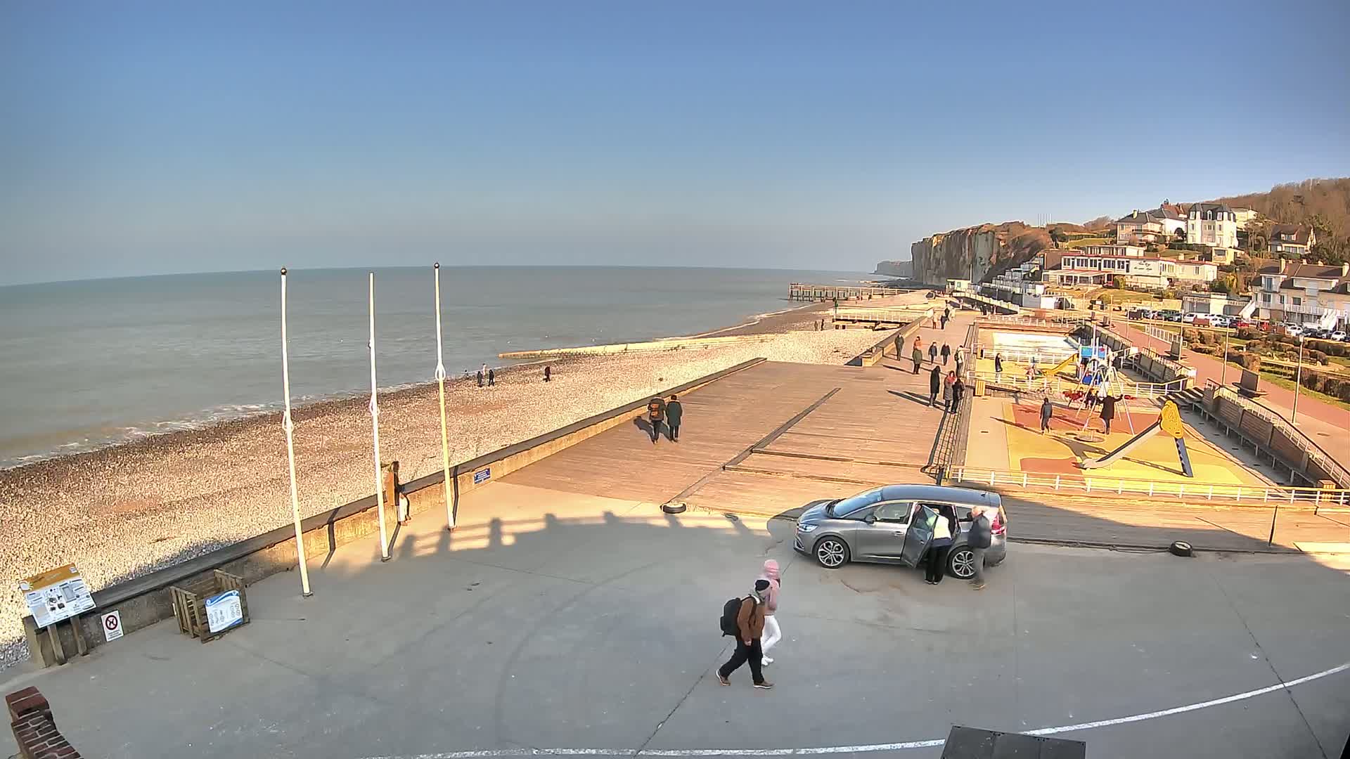 A sunny day at a pebble beach with a wooden boardwalk, playground, and a few people walking along the shore near a seaside cliff.