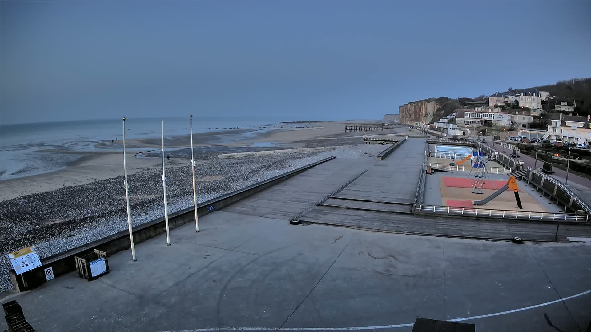 A mostly empty beach with a playground and a boardwalk overlooks a calm sea under a clear, dusky sky.