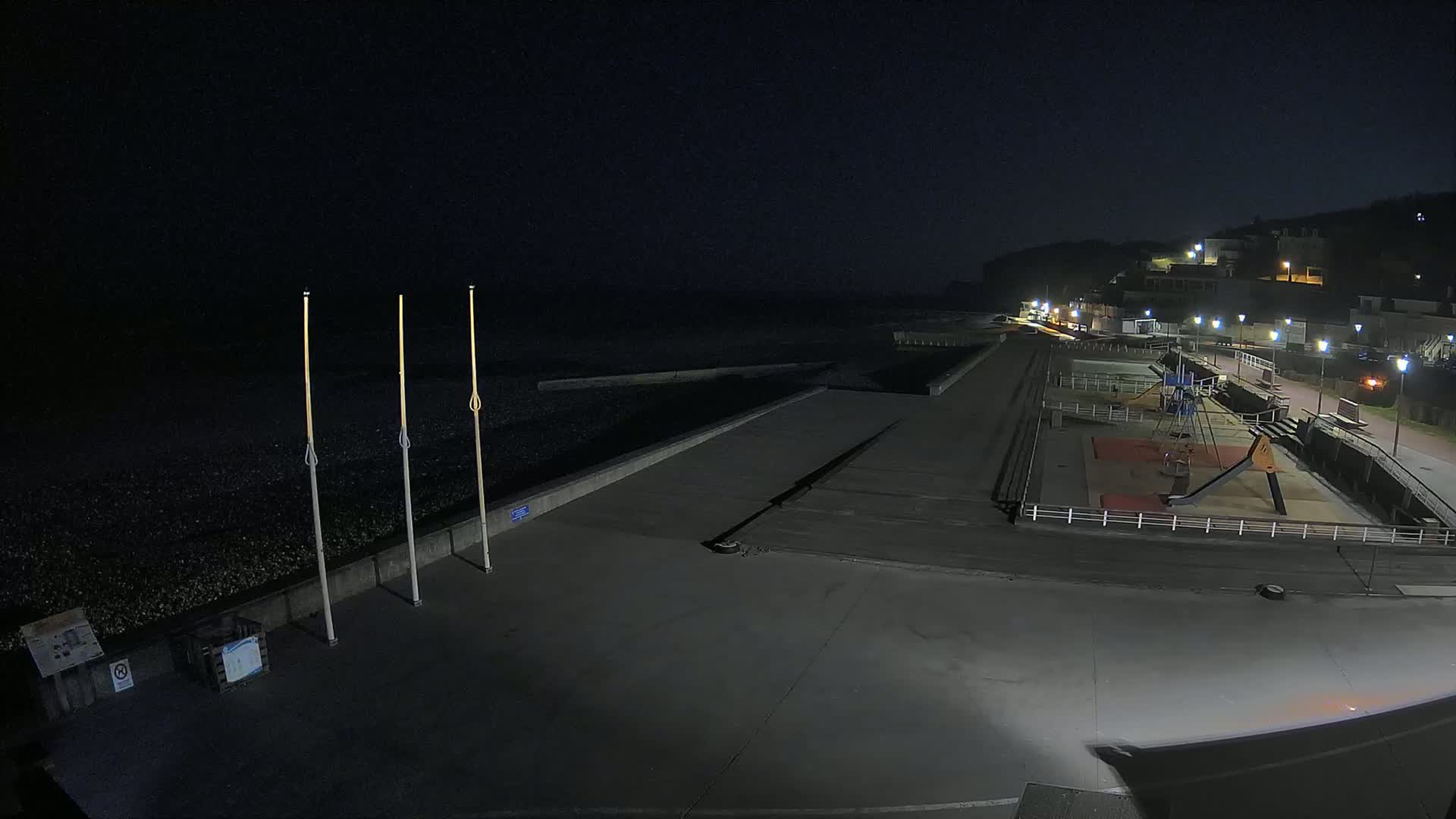 A nighttime view of a mostly empty seaside promenade with a playground and a dark, pebble beach, under a clear sky.