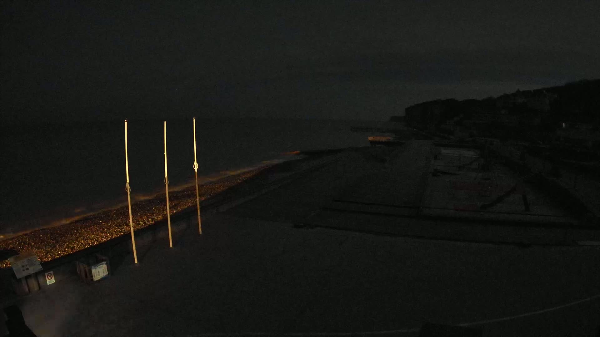 Three tall, slender, lit posts stand on a dark beach at night near a dark body of water under a dark, cloudy sky.