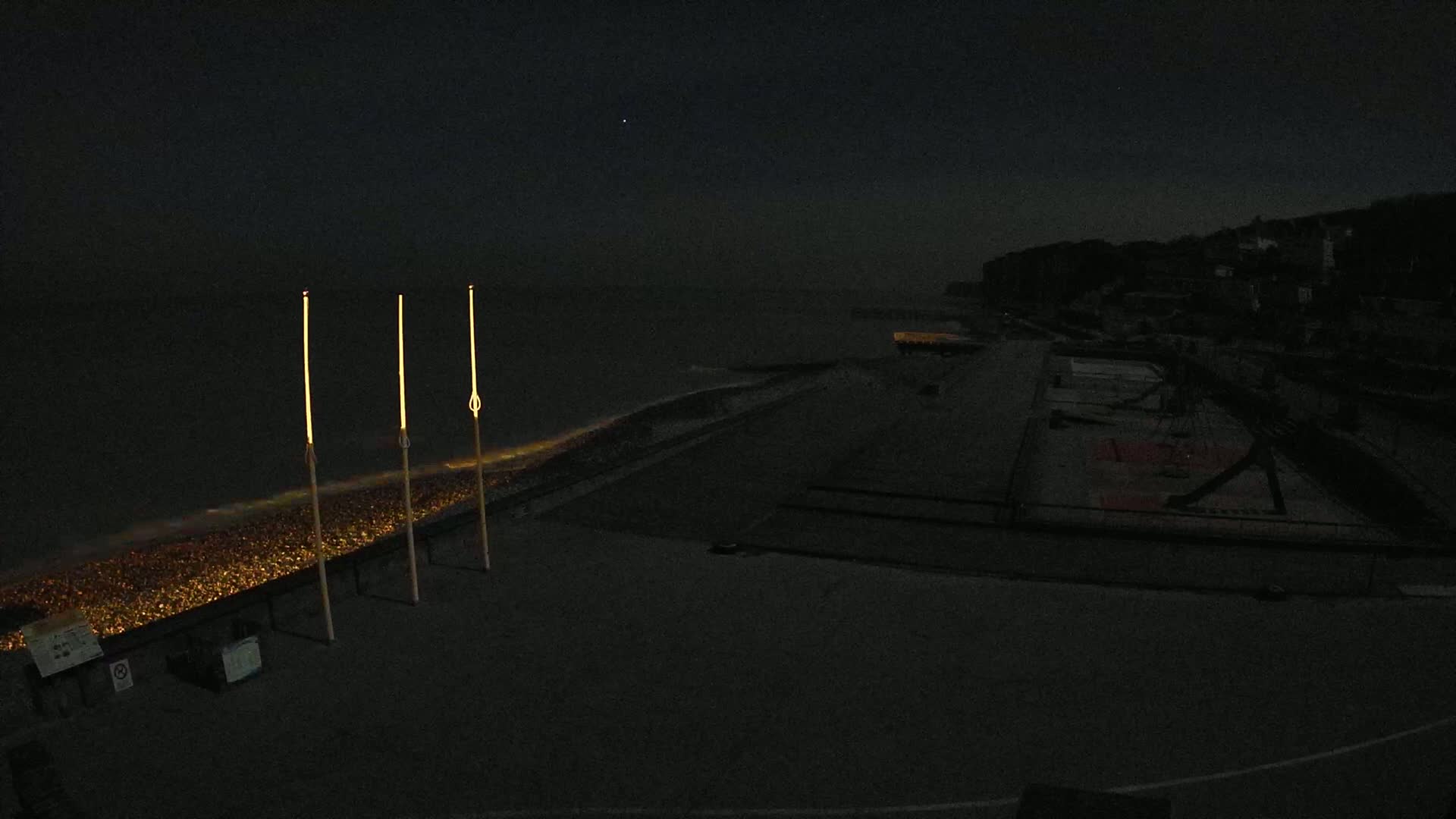 At night, three tall, illuminated poles stand near a dark, pebble-covered shoreline next to a dark body of water under a mostly clear sky.