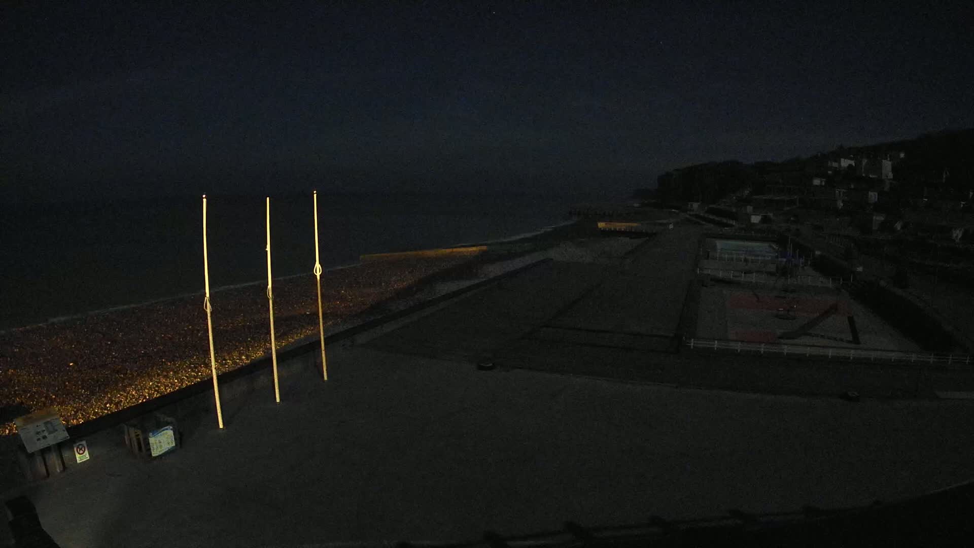 At night, three tall, thin poles stand illuminated near a dark, pebble-covered beach next to a mostly empty playground and a dark sea under a clear, starlit sky.