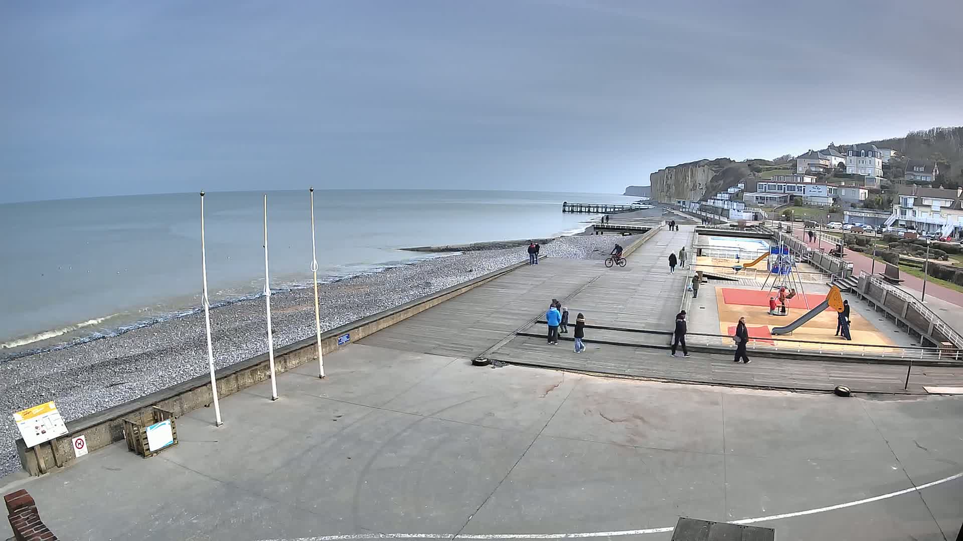 A mostly empty seaside promenade with a playground, a pier, and white cliffs in the background under a cloudy sky.