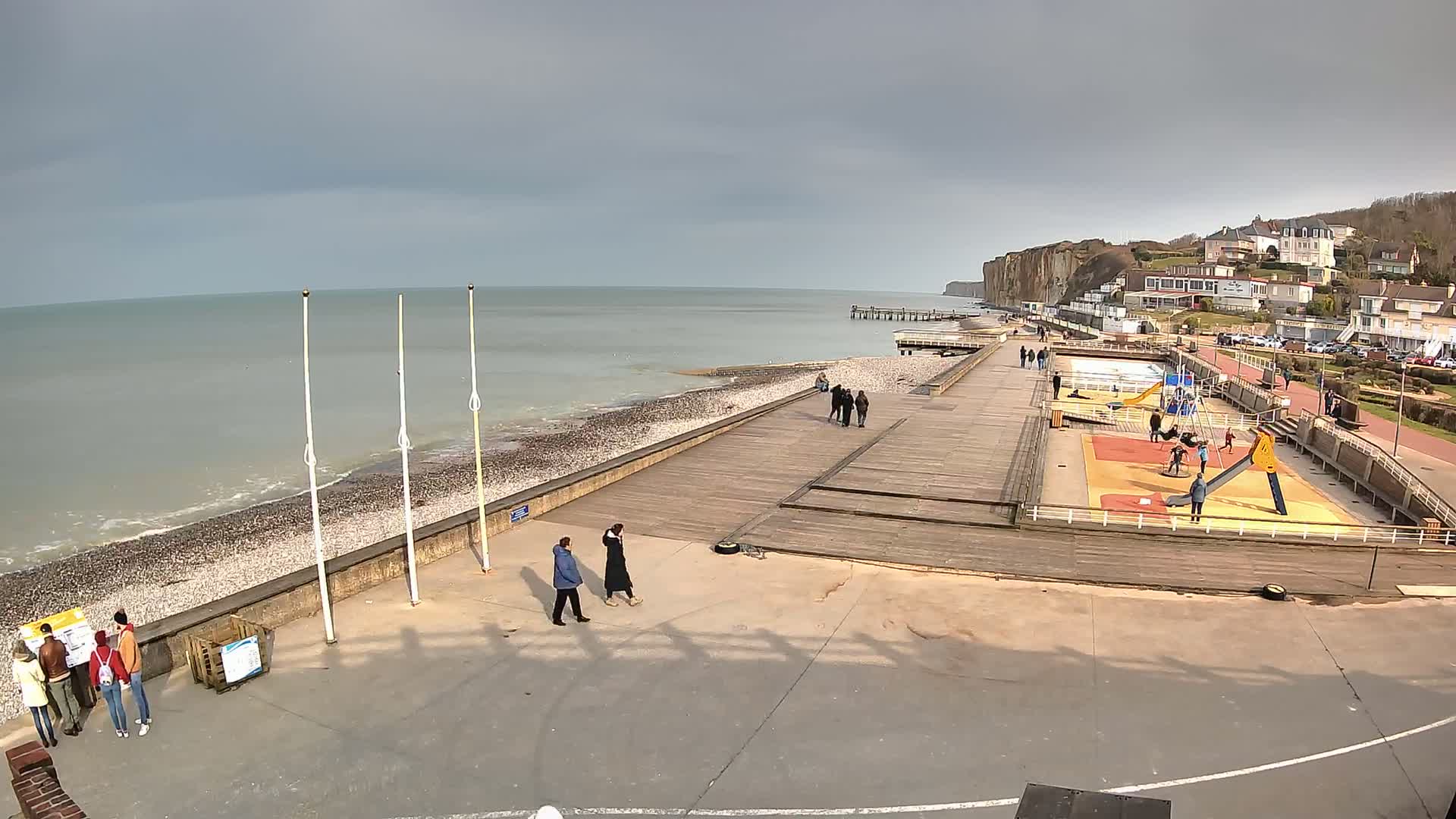 A mostly cloudy day shows a seaside boardwalk with a playground and a few people walking near a pebble beach and white cliffs in the distance.