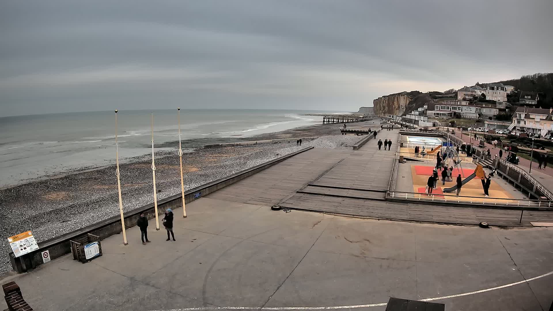 A pebble beach with a wooden boardwalk and playground is seen under an overcast sky, next to white-capped waves and white buildings on a cliff.