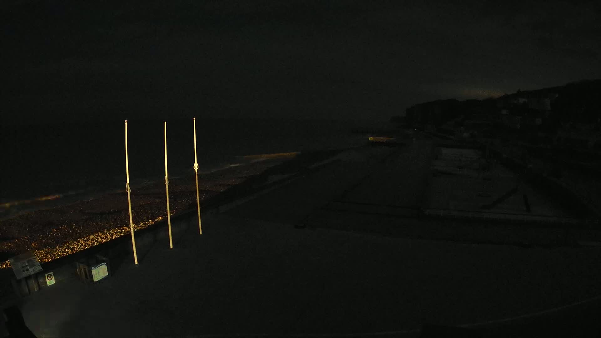 Three tall, slender lights stand illuminated on a dark beach at night, overlooking a dimly lit shoreline and a dark, silhouetted hill in the distance under a cloudy sky.