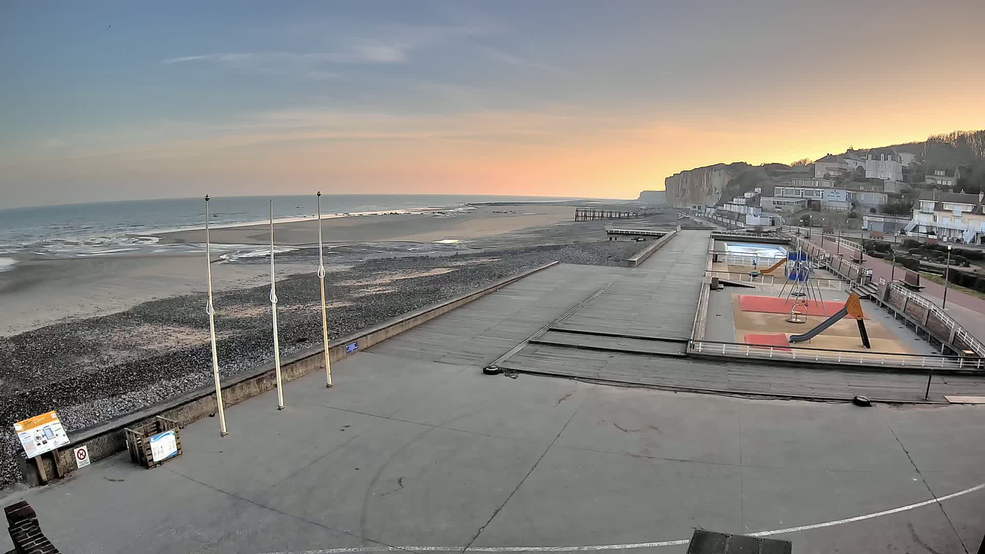 A seaside playground and promenade overlook a calm ocean beach at sunset, with white cliffs in the background.