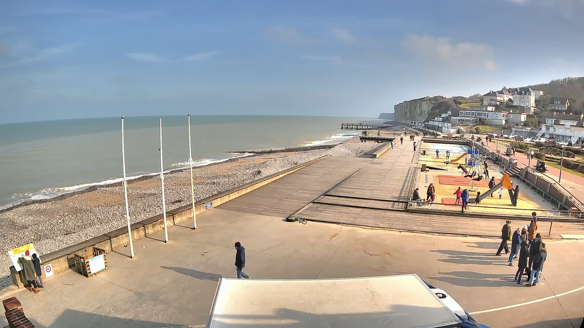 A seaside boardwalk with a playground and people, overlooking a rocky beach and calm ocean under a sunny sky.