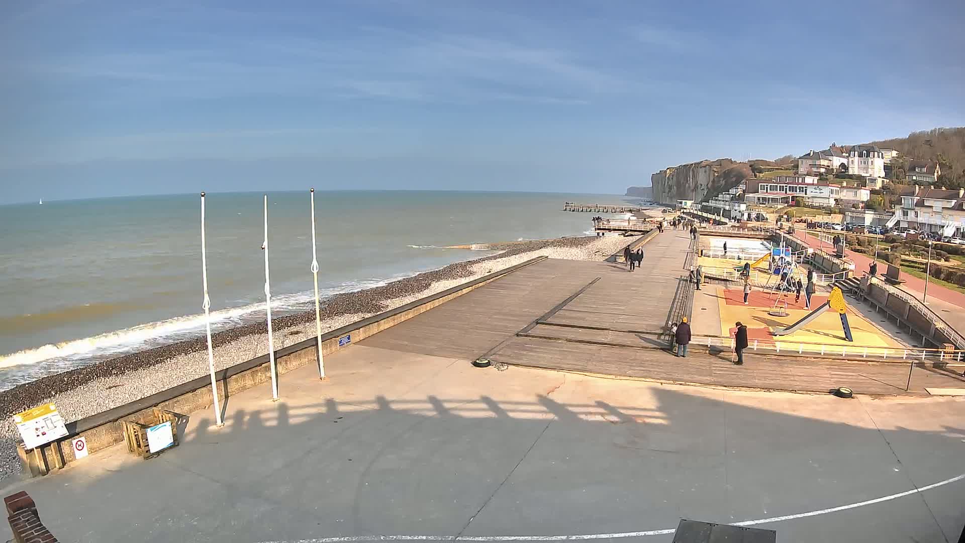A seaside boardwalk with a playground and people, overlooking a rocky beach and ocean waves under a clear, sunny sky.