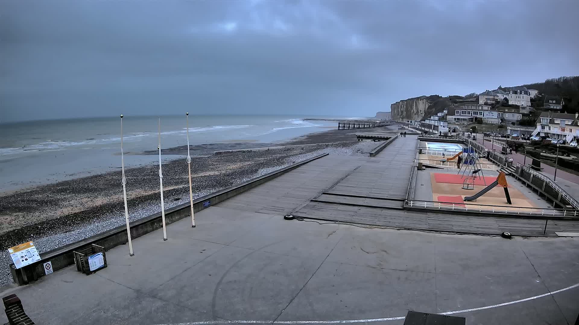 A mostly empty beach and boardwalk area with a small playground and a pool, under an overcast sky with a gray sea and white capped waves.