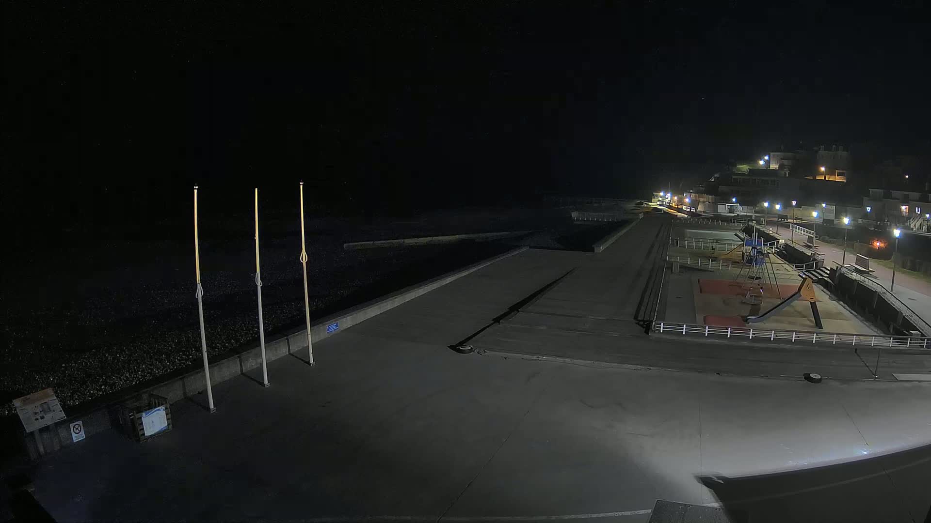 A nighttime, overhead view shows an empty playground and paved beachfront area next to a dark, pebble-covered shoreline, illuminated by streetlights.