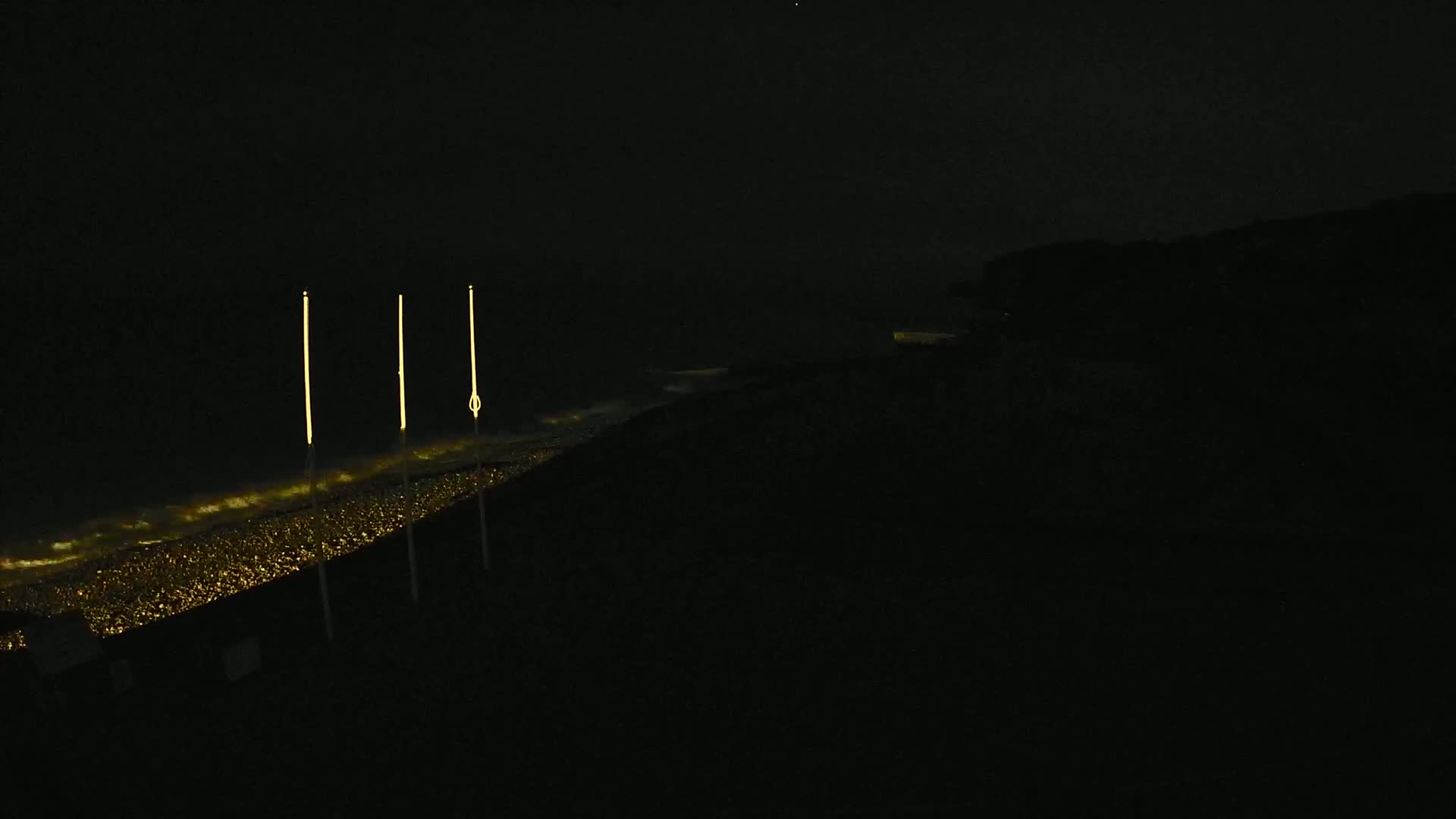 Three tall, thin, illuminated structures stand on a dark shoreline at night, with a faintly lit ocean in the foreground.