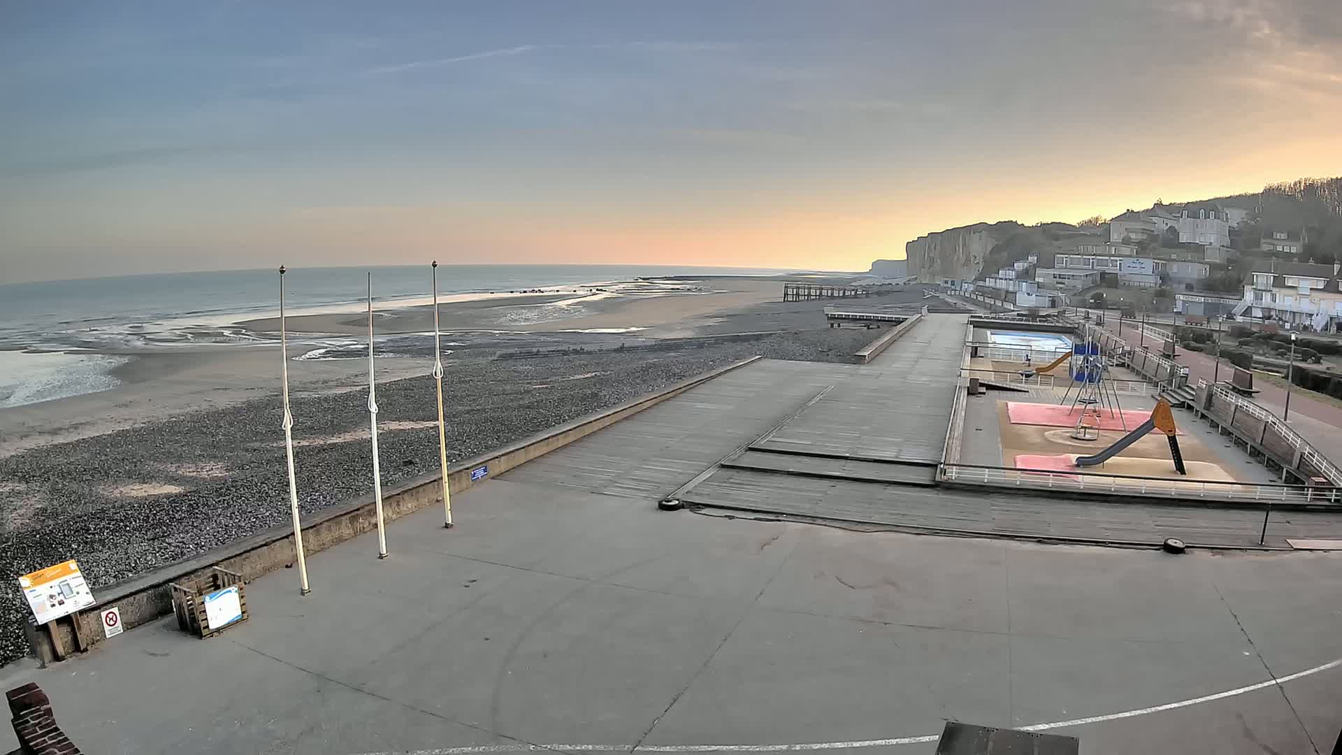 A beach with a playground and a boardwalk is seen at sunset, overlooking a calm ocean and white cliffs.