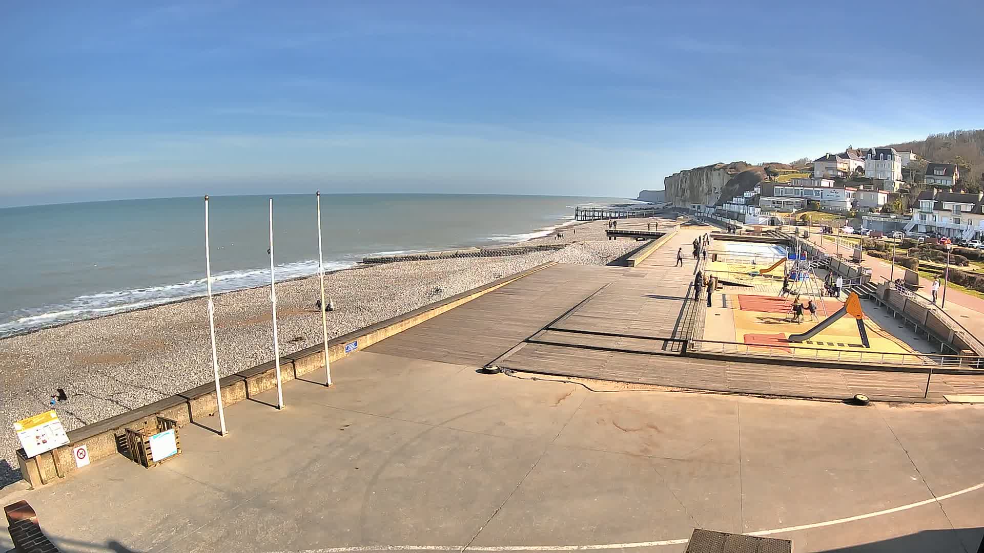 A pebble beach with a wooden boardwalk and playground is adjacent to white cliffs and a calm ocean under a clear blue sky.