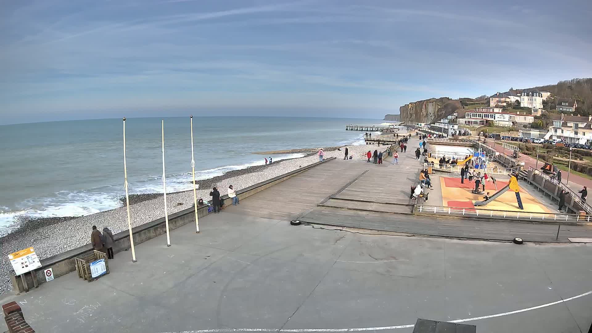 A seaside boardwalk with a playground and people, under a partly cloudy sky, overlooks a rocky beach and the ocean with small waves.
