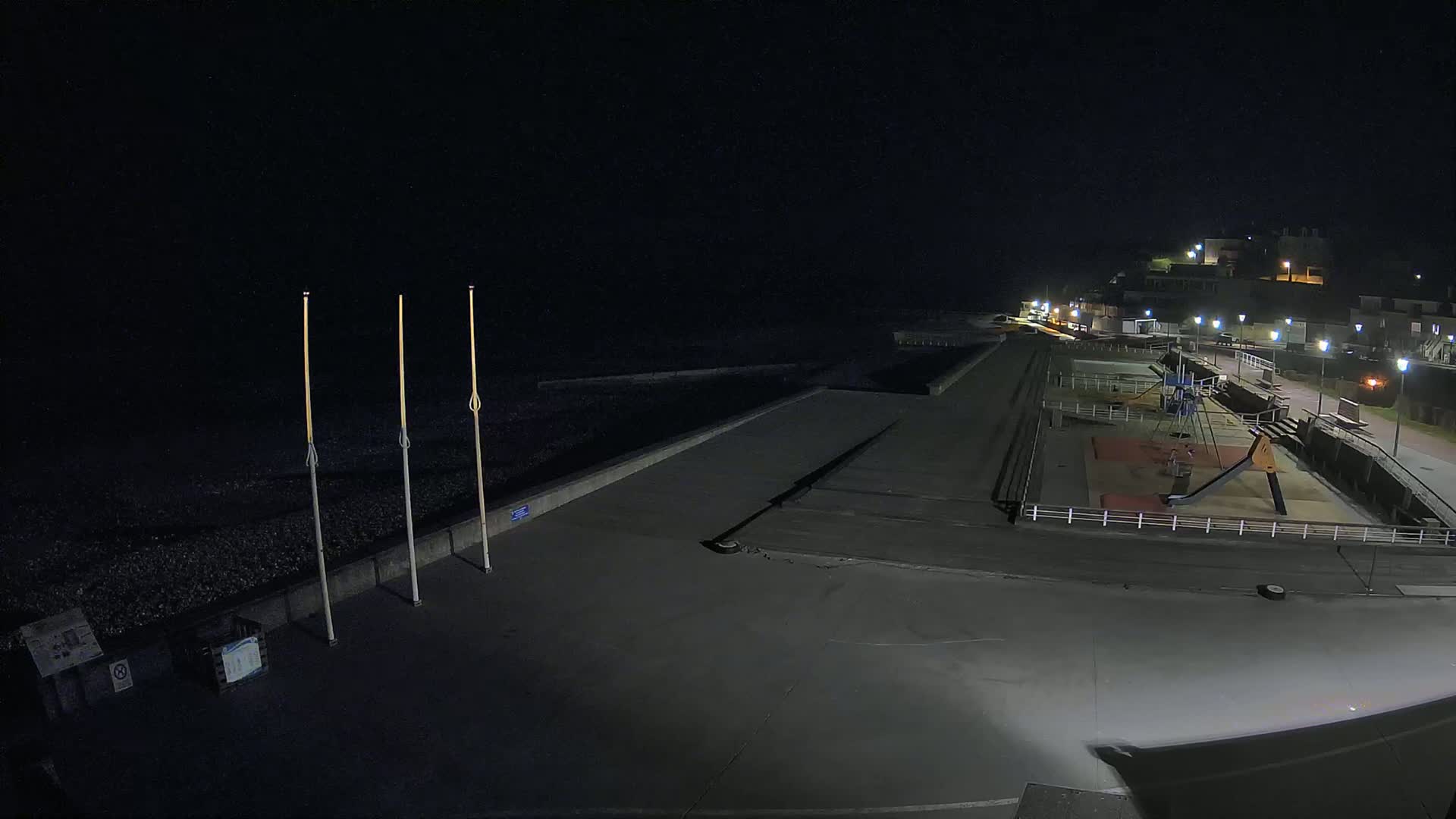 A nighttime, seaside view of an empty paved area with a playground and a dark beach beyond a low seawall, illuminated by artificial lights.