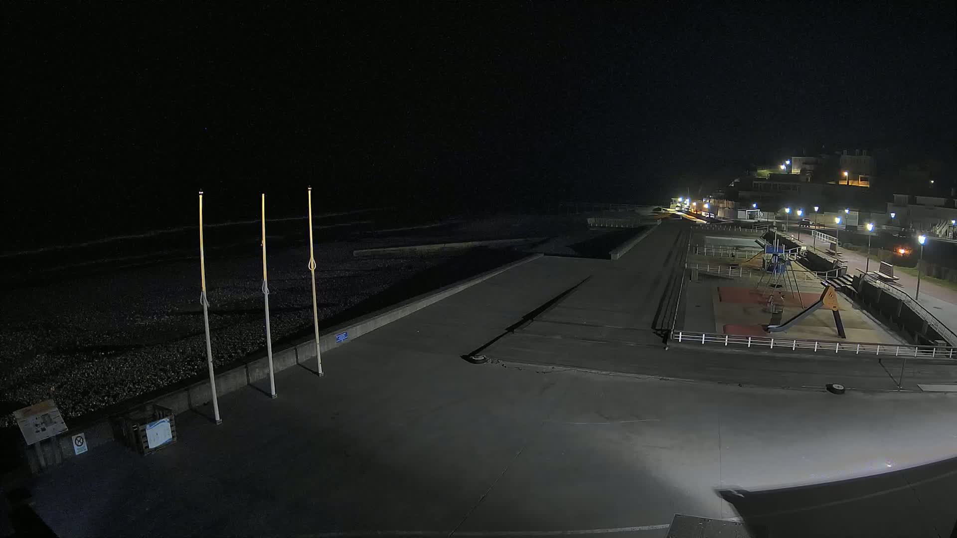 A nighttime view of a mostly empty beachfront area with a playground, a seawall, and a dark ocean under a clear sky.